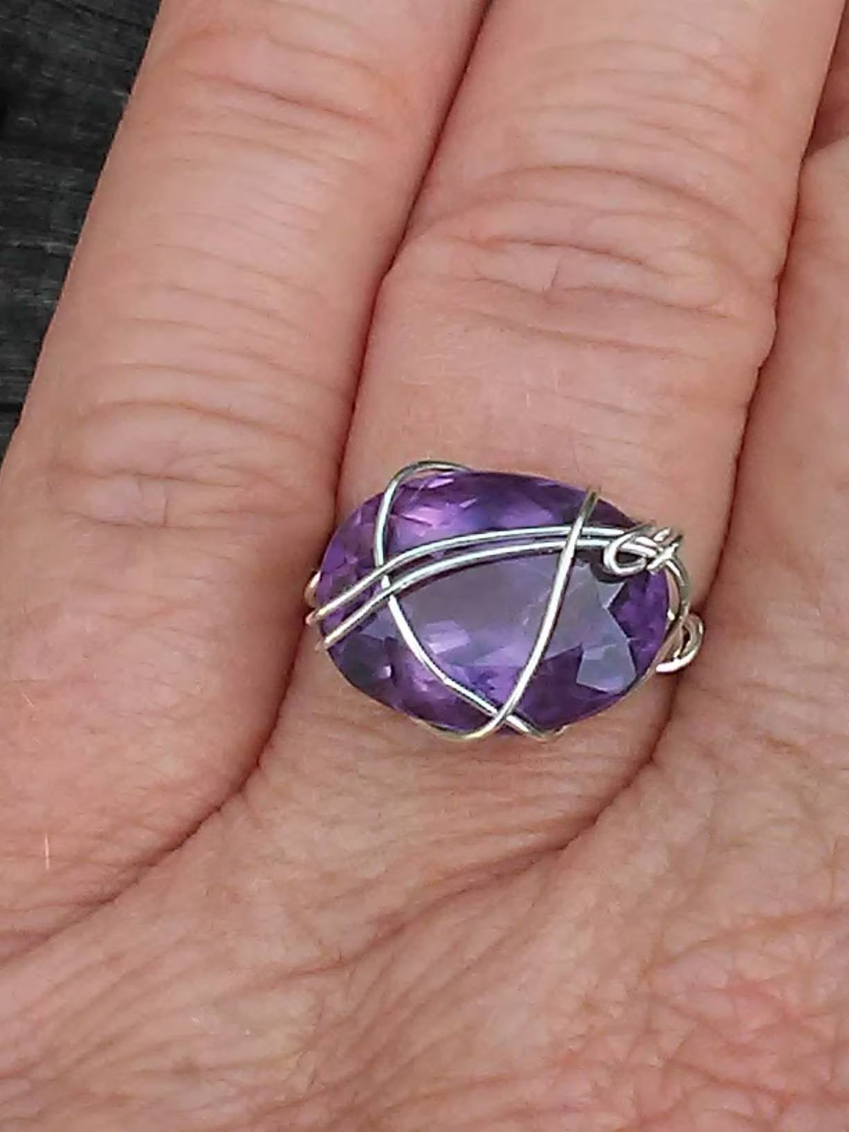 A silver wire-wrapped ring with a faceted purple gemstone, shown worn on a person's finger.