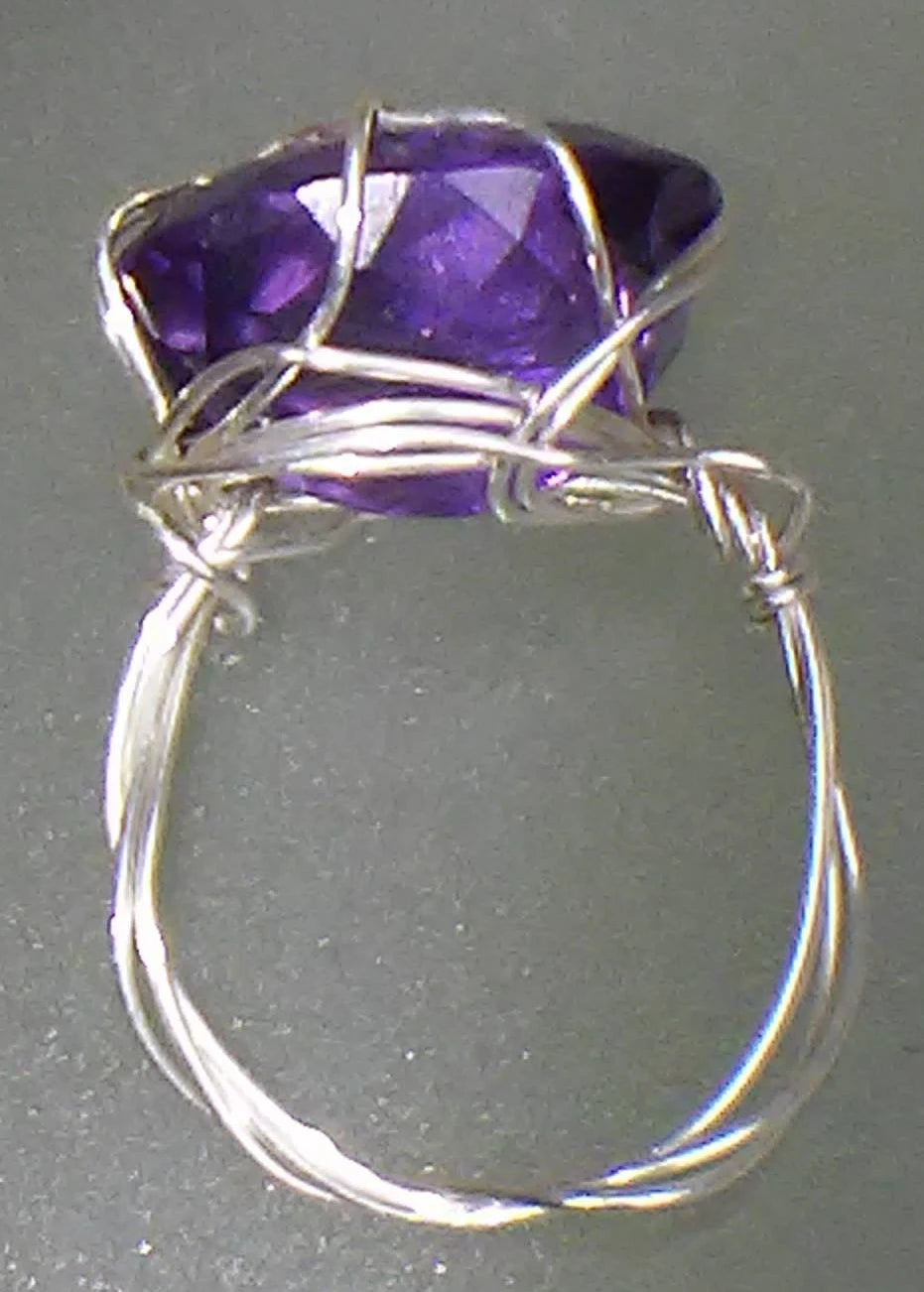 A handmade silver wire-wrapped ring featuring a large, faceted purple gemstone, shown against a dark gray background.