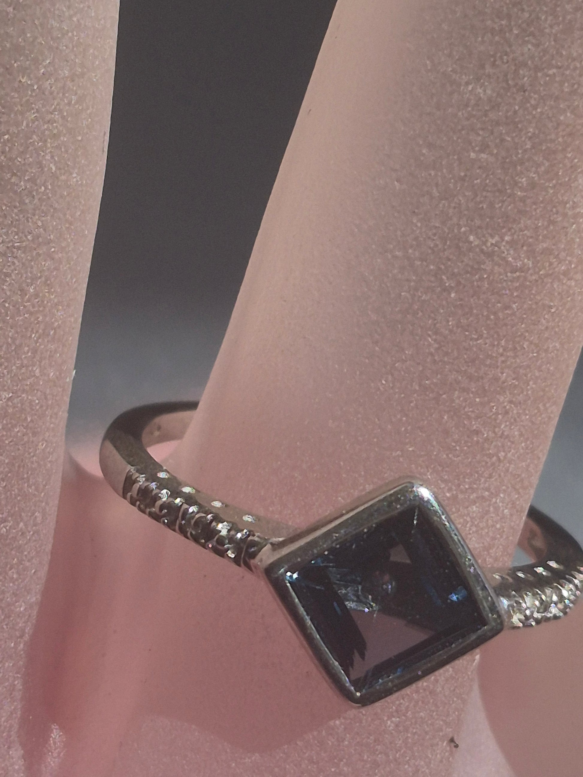 A silver ring with a square-cut dark blue gemstone and textured band details, displayed on a pink ring stand.