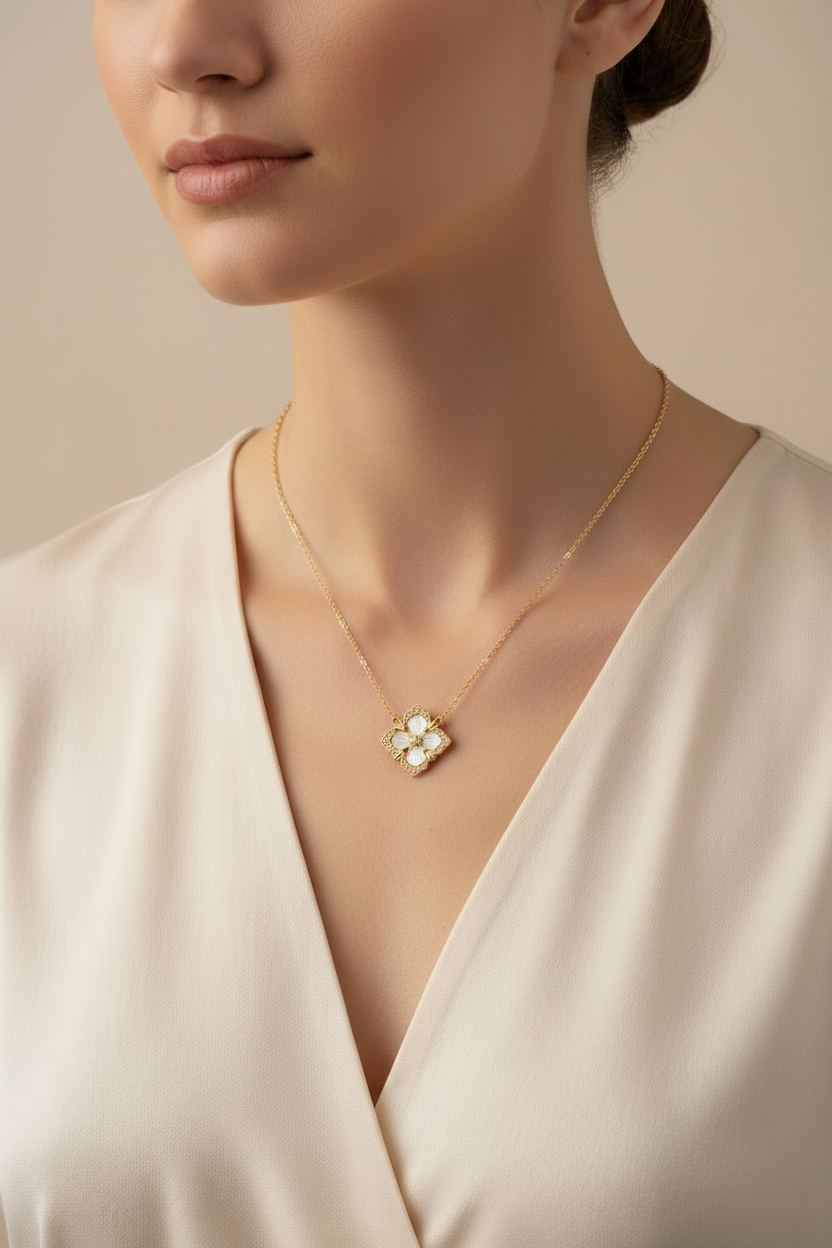 A close-up of a model wearing a delicate gold necklace with a four-petal flower pendant featuring white pearl-like accents, paired with a cream-colored satin top against a soft beige background.