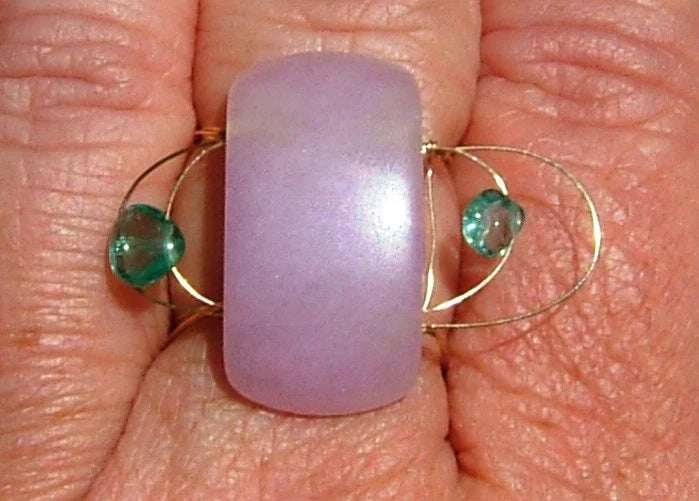 A close-up of a lavender purple rectangular cabochon stone set in a gold ring with two small green faceted beads, shown resting on a person's fingers.