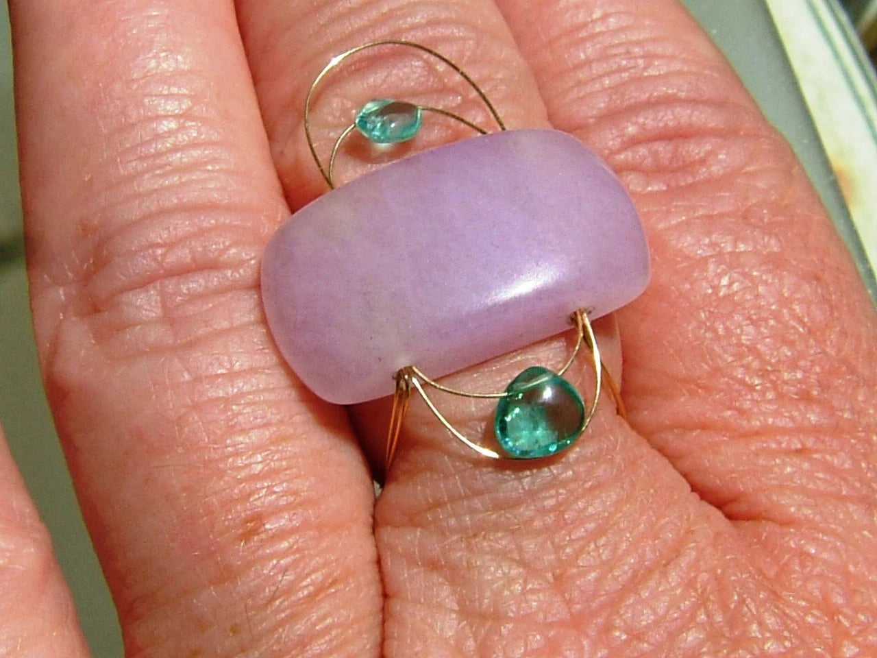A close-up of a hand wearing a unique ring featuring a large, smooth lavender-purple cabochon stone set in gold-toned metal with two small green teardrop gemstones.