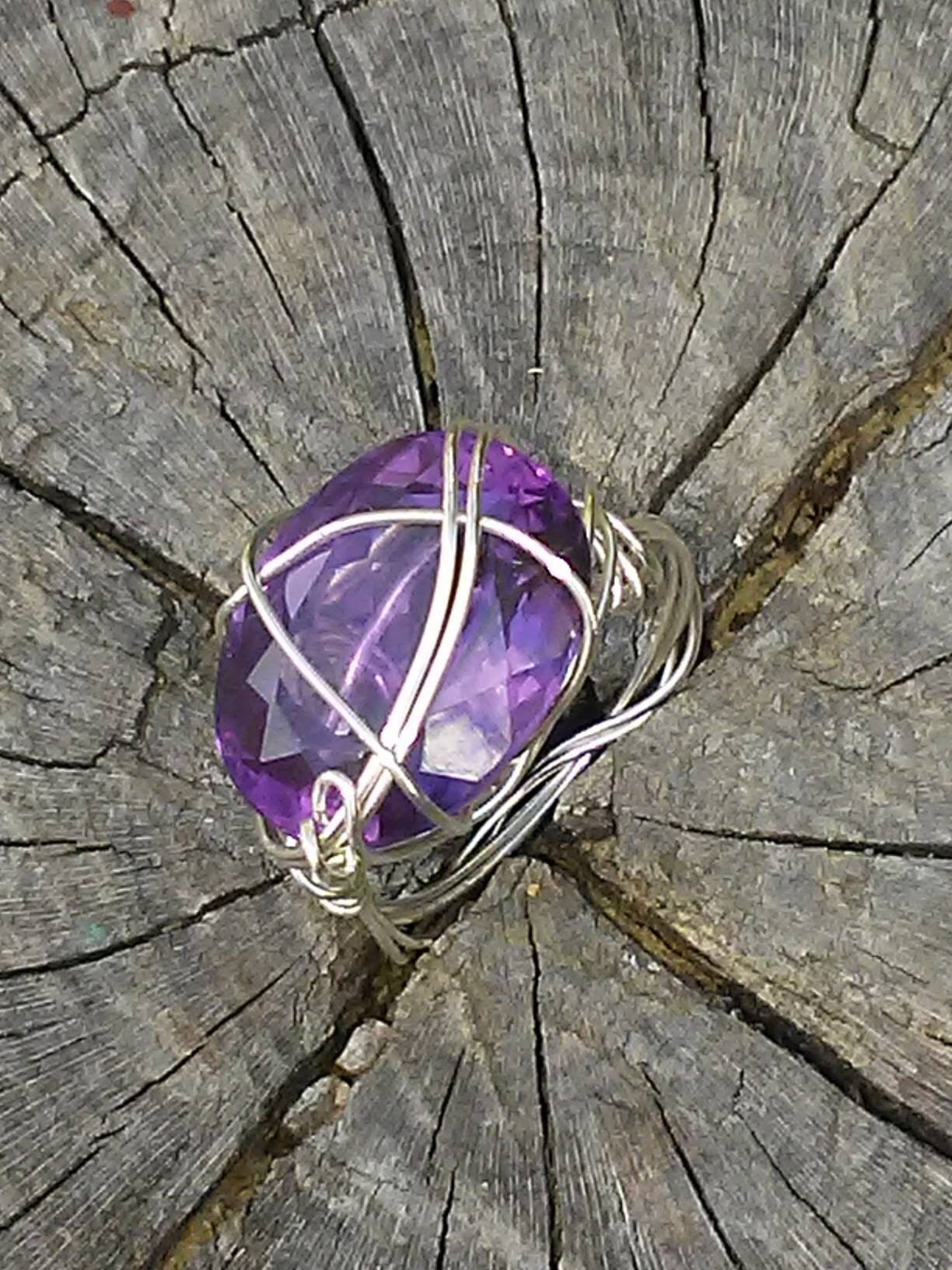 A faceted purple gemstone ring wrapped in silver-toned wire, displayed on a weathered tree stump.