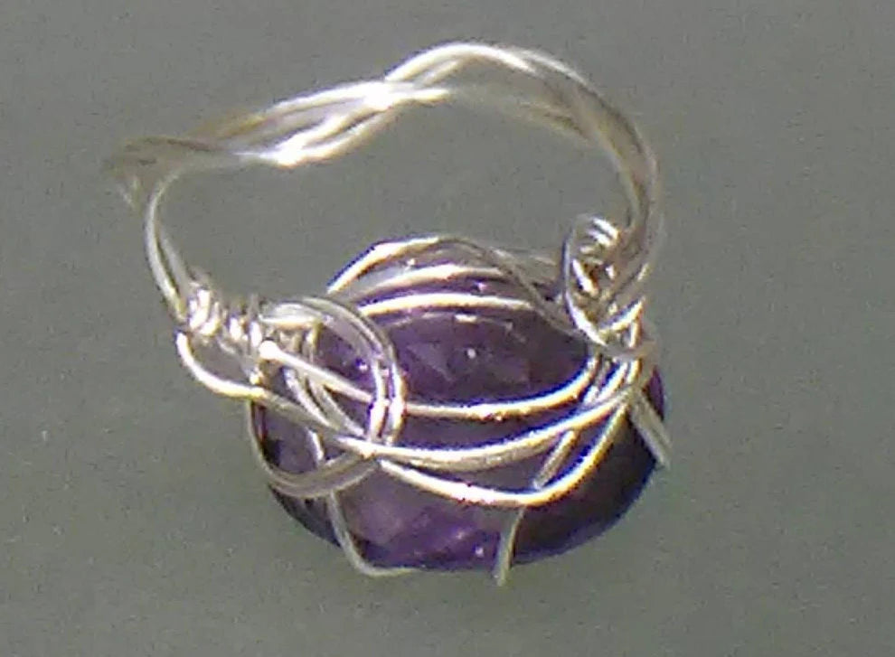 A handmade silver wire ring featuring a twisted band and a central cluster of silver wires wrapping around a faceted purple gemstone, set against a plain gray background.