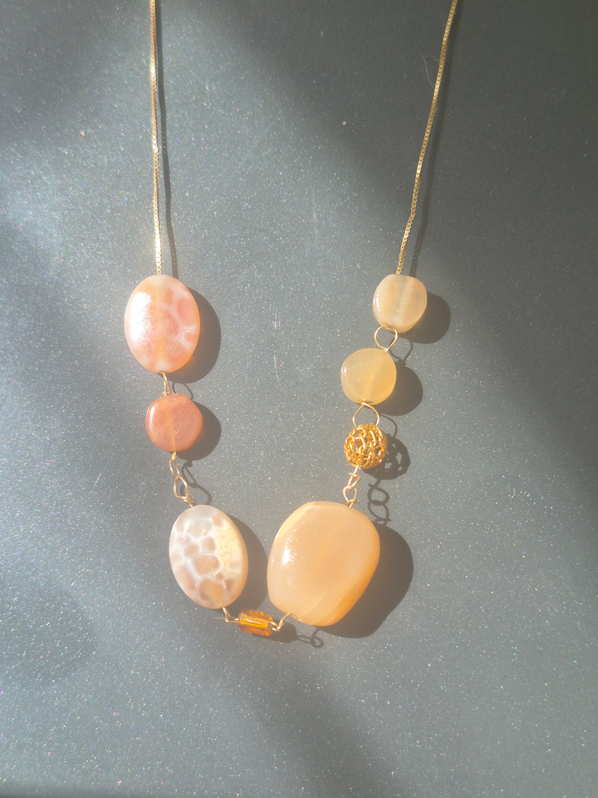 A handmade beaded necklace featuring polished peach, orange, and cream-colored stones of various shapes, accented by a small gold wire bead and a tiny amber accent, displayed on a dark gray surface with dramatic sunlight and shadows.