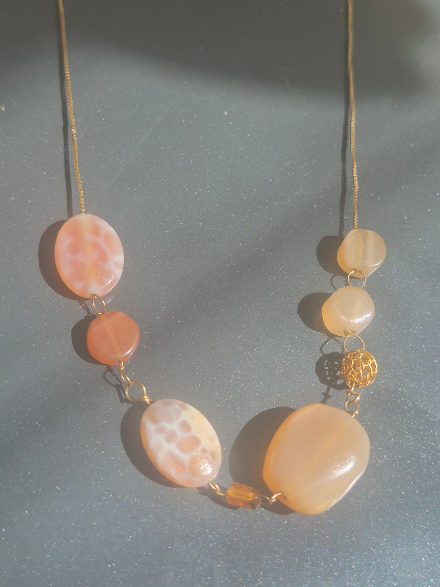 A gold-tone necklace featuring large, organic-shaped peach and cream agate beads with natural marbled patterns, accented by a small gold bead and a gold filigree spacer, displayed on a textured gray surface.