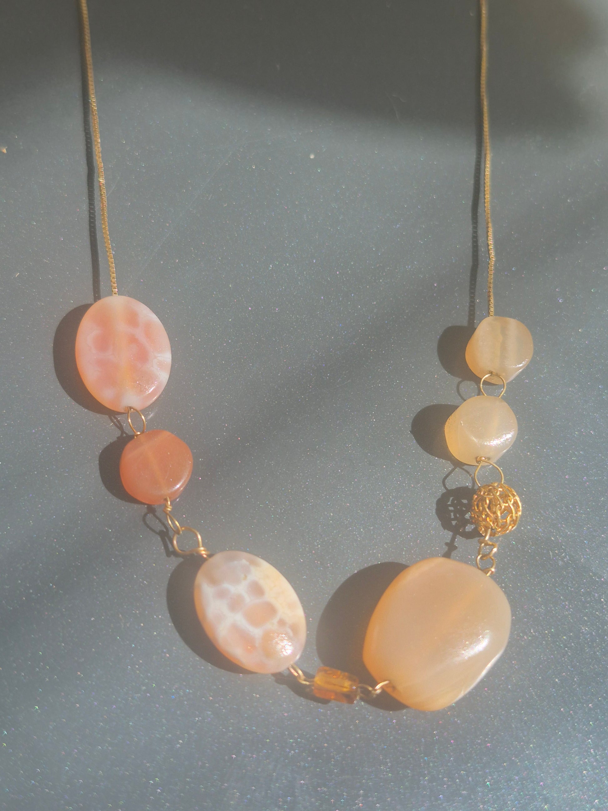 A gold-tone necklace featuring large, organic-shaped peach and cream agate beads with natural marbled patterns, accented by a small gold bead and a gold filigree spacer, displayed on a textured gray surface.