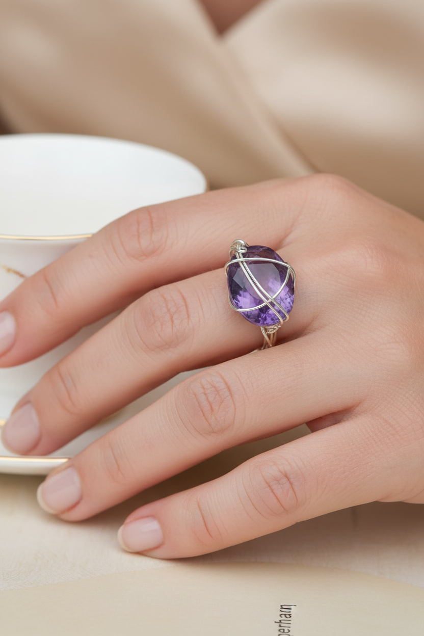 A close-up of a hand wearing a silver ring with a faceted purple gemstone, set against a soft beige background with a white teacup partially visible.