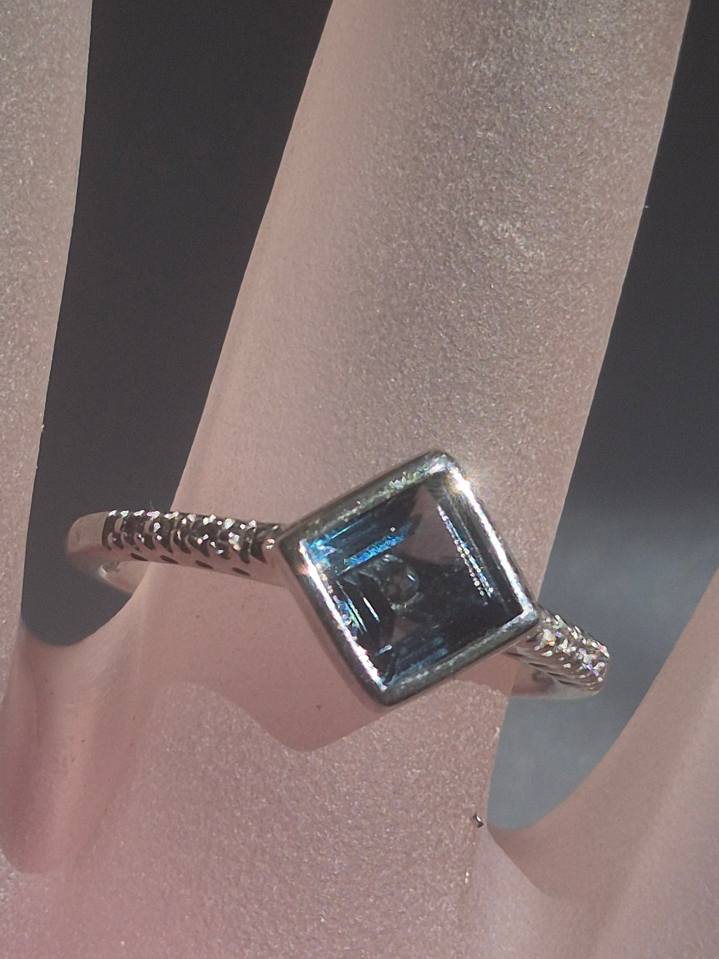 A silver ring with a square-cut light blue gemstone, displayed on a pink ring stand.