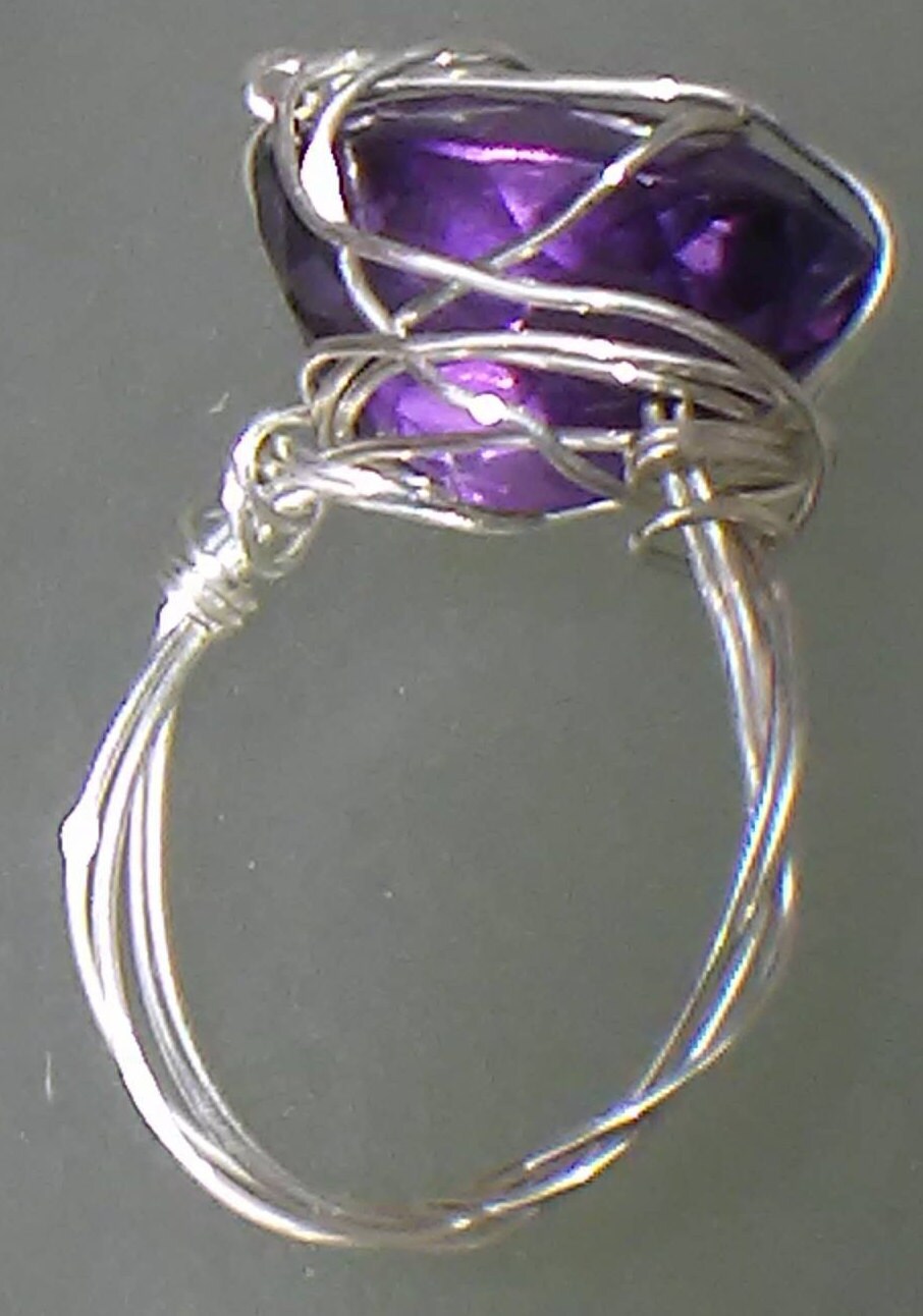A silver wire-wrapped ring featuring a large, faceted purple amethyst gemstone, shown against a neutral gray background.